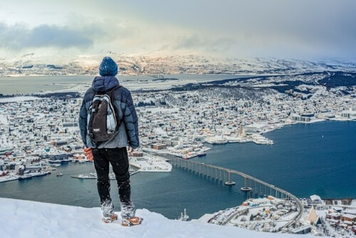 山頂から雪景色のトロムソ市街や雪山、フィヨルドを眺める男性